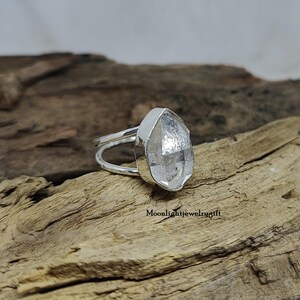 May include: A silver ring with a clear, faceted quartz stone. The stone is set in a bezel setting and the ring band is simple and delicate.