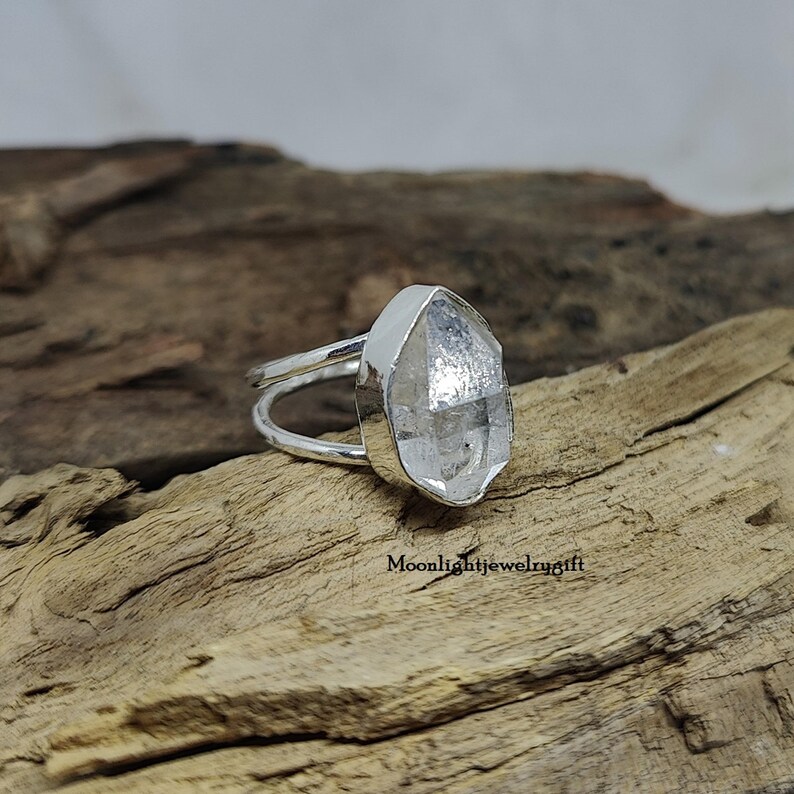 May include: A silver ring with a clear, faceted quartz stone. The stone is set in a bezel setting and the ring band is simple and delicate.