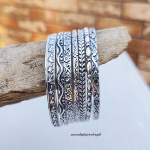 Pu&ograve; includere: Un set di otto braccialetti d'argento con intricati disegni in rilievo. I braccialetti sono impilati su un pezzo di legno galleggiante.