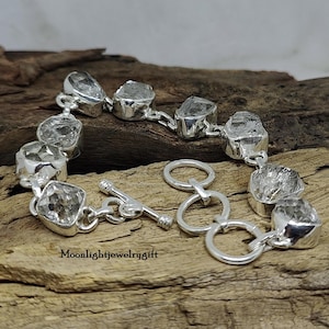May include: A silver bracelet with a toggle clasp features clear, faceted, rough-cut gemstones. The bracelet is on a wooden surface.