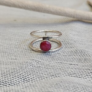 Może przedstawiać: Srebrny pierścionek z podw&oacute;jnym obramowaniem, w środku kt&oacute;rego znajduje się okrągły, czerwony kamień szlachetny.