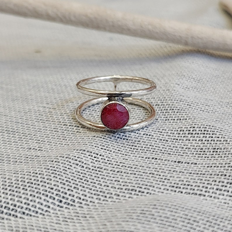 Może przedstawiać: Srebrny pierścionek z podw&oacute;jnym obramowaniem, w środku kt&oacute;rego znajduje się okrągły, czerwony kamień szlachetny.