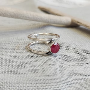 Może przedstawiać: Srebrny pierścionek z podw&oacute;jną obręczą i czerwonym kamieniem szlachetnym. Pierścionek leży na białym tle z tkaniny.