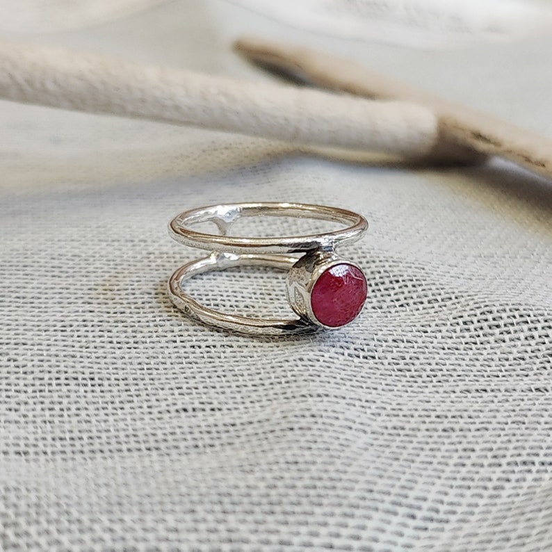 Może przedstawiać: Srebrny pierścionek z podw&oacute;jną obręczą i czerwonym kamieniem szlachetnym. Pierścionek leży na białym tle z tkaniny.