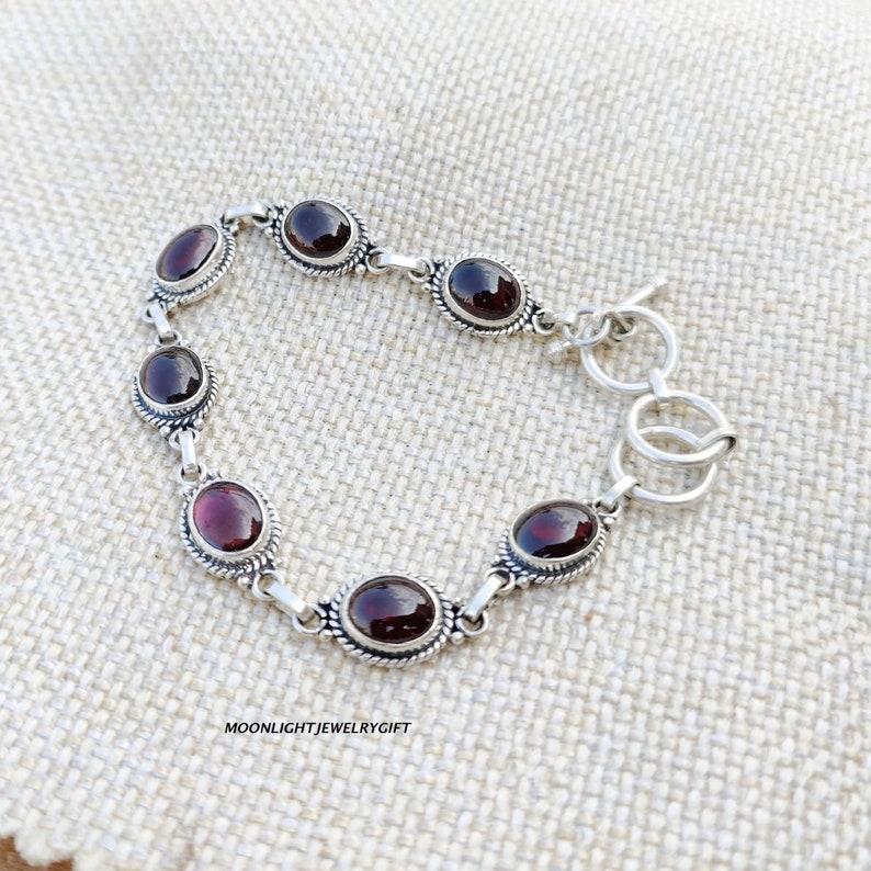 K&ouml;nnte beinhalten: Ein silbernes Armband mit einem Schiebeverschluss verf&uuml;gt &uuml;ber sieben ovale rote Edelsteine. Das Armband liegt auf einem beigefarbenen Stoffhintergrund.