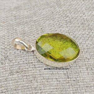 May include: A silver pendant with a large, faceted, oval-shaped green gemstone. The pendant is on a textured, light gray surface.  Moonlightjewelrygift is printed below the pendant.