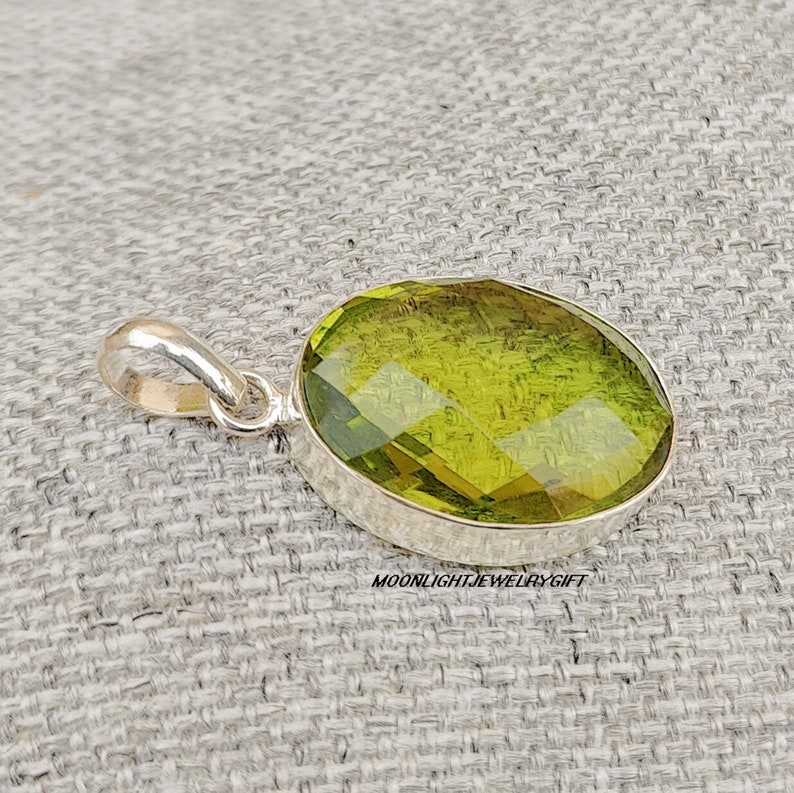 May include: A silver pendant with a large, faceted, oval-shaped green gemstone. The pendant is on a textured, light gray surface.  Moonlightjewelrygift is printed below the pendant.