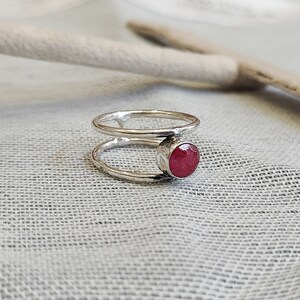 Może przedstawiać: Srebrny pierścionek z podw&oacute;jną obręczą i czerwonym kamieniem szlachetnym. Pierścionek leży na białym tle z tkaniny.