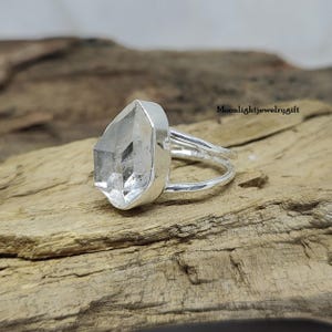 May include: A silver ring with a clear quartz gemstone. The ring is set on a wooden surface.