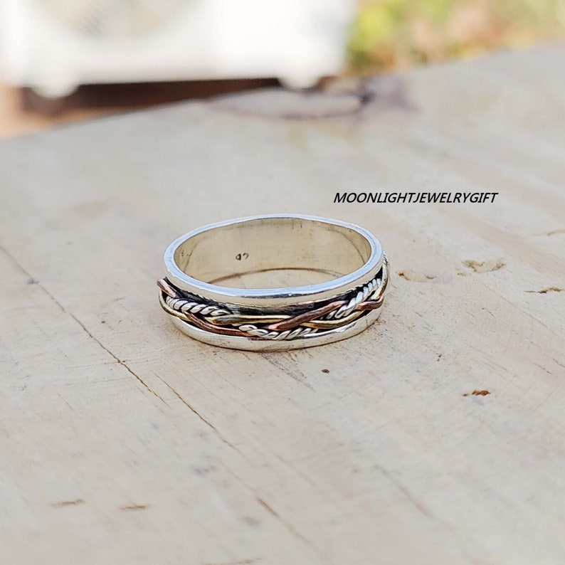 Pu&ograve; includere: Un anello a fascia d'argento con un design intrecciato in rame e filo d'argento. L'anello &egrave; su una superficie di legno.