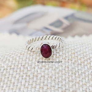 May include: A silver ring with a twisted band and a red oval gemstone. The ring is on a white fabric background.