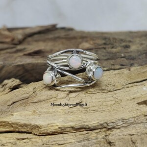 Pu&ograve; includere: Un anello d'argento con tre pietre di opale bianco incastonate in un design a fascia attorcigliata. L'anello &egrave; su una superficie di legno.
