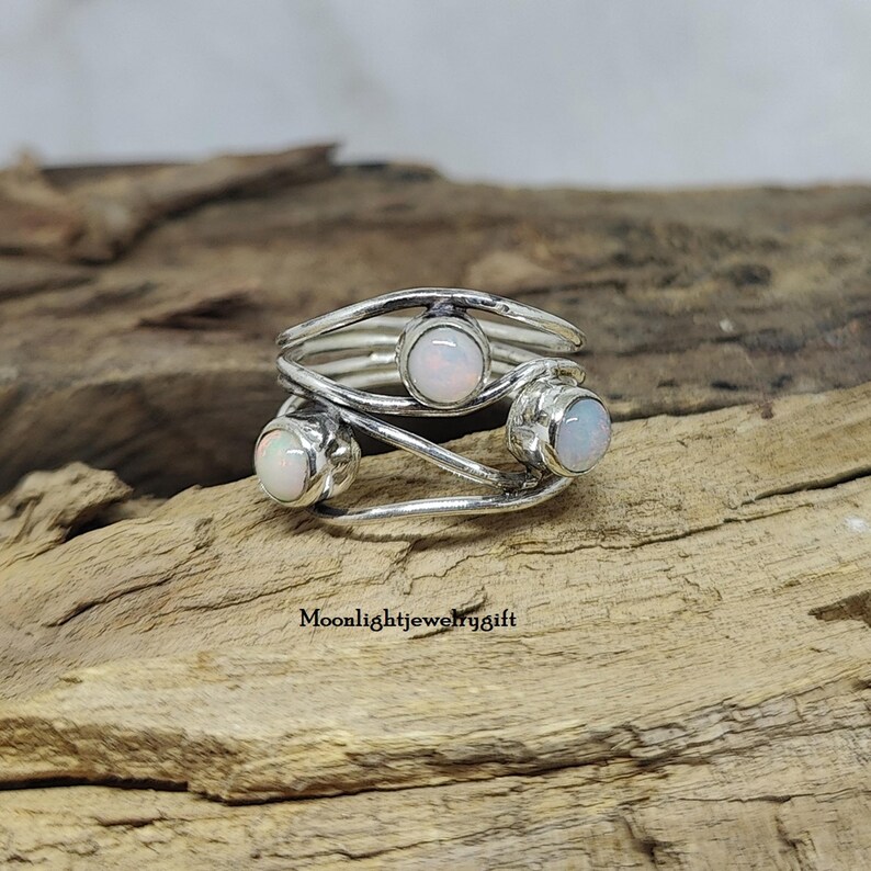 Pu&ograve; includere: Un anello d'argento con tre pietre di opale bianco incastonate in un design a fascia attorcigliata. L'anello &egrave; su una superficie di legno.