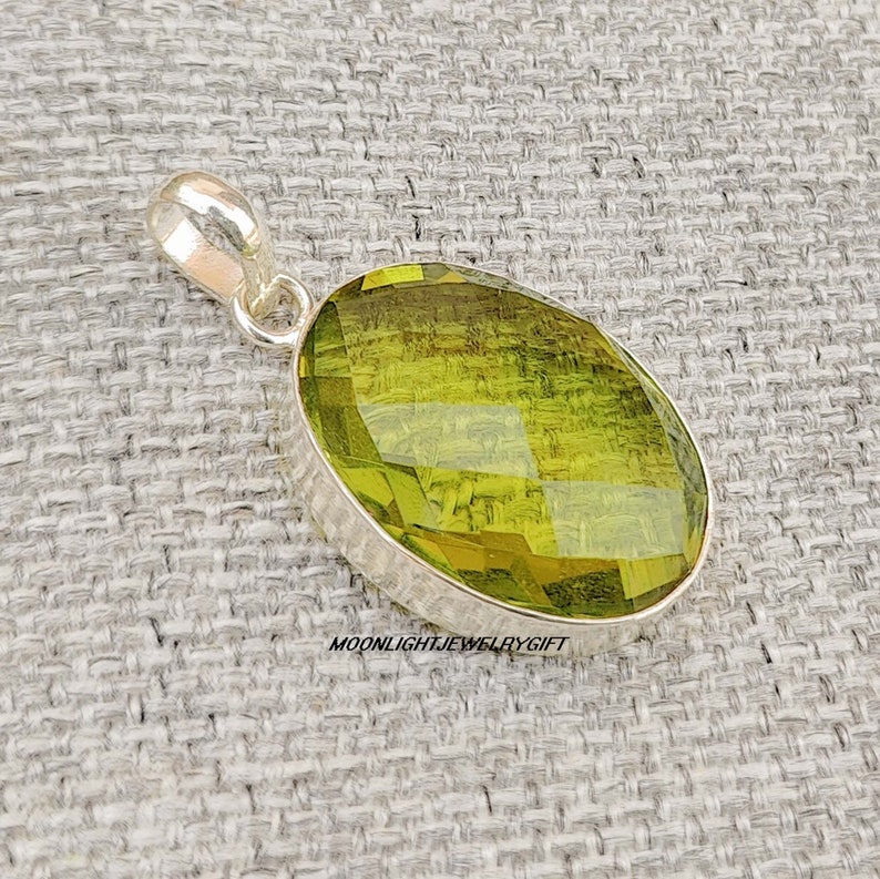 May include: A silver pendant with a large, oval-shaped, faceted green gemstone. The pendant is on a textured, light gray surface.