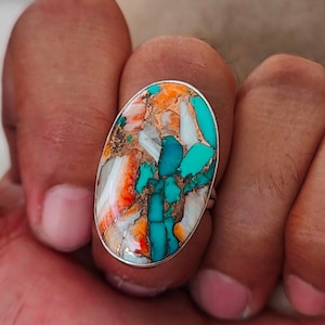 May include: Oval-shaped ring featuring a vibrant, multicolored stone. The stone showcases a mosaic of turquoise, orange, white, and copper hues, set in a silver band. The ring's design is eye-catching and suitable for everyday wear or special occasions.