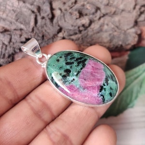 Puede incluir: Colgante ovalado con una piedra preciosa de rubí zoisita pulida. La piedra presenta una vibrante mezcla de colores rosa, verde y negro, engastada en un bisel plateado. El colgante tiene una argolla plateada.