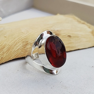 May include: A silver ring with a large, oval, deep red gemstone. The ring has a modern design with a wide band and open sides. The gemstone is smooth and polished, reflecting light.