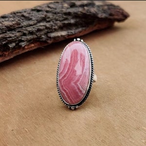 May include: Oval-shaped ring featuring a large, pink gemstone with white veining, set in a silver-toned metal frame. The ring has a twisted rope-like border and is displayed on a wooden surface.