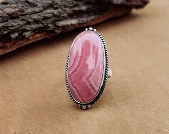 May include: Oval-shaped ring featuring a large, pink gemstone with white veining, set in a silver-toned metal frame. The ring has a twisted rope-like border and is displayed on a wooden surface.