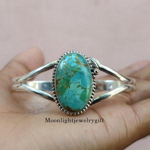May include: A silver cuff bracelet featuring a large, oval-shaped turquoise stone with brown matrix veining. The stone is set in a beaded silver bezel with a small leaf detail. The band is split into two polished silver arms. Text reads Moonlightjewelrygift.