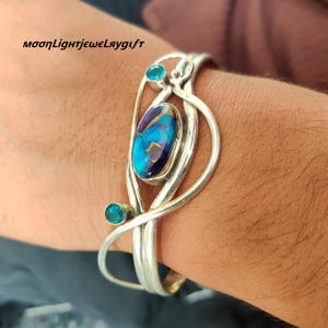 May include: A silver cuff bracelet featuring a central oval-shaped blue and purple stone with copper veining, accented by two smaller round blue stones. The metal frame consists of smooth, curved silver wires that wrap around the wrist.