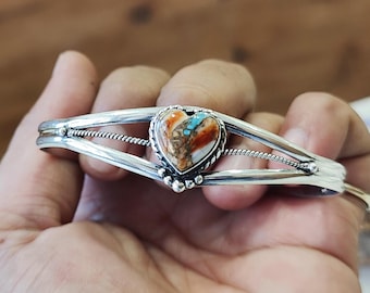 May include: A silver bracelet with a heart-shaped turquoise stone set in the center. The bracelet has a delicate design with two bands and a twisted wire detail.
