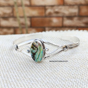 Puede incluir: Pulsera de brazalete de plata con una incrustación ovalada de concha de abulón. La pulsera tiene un diseño delicado y curvo con pequeños acentos.