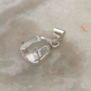 May include: A clear quartz crystal pendant with a silver setting. The crystal is faceted and has a natural, rough appearance. The pendant is attached to a silver bail.