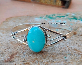 Könnte beinhalten: Ein silbernes Armreif-Armband mit einem großen ovalen Türkis-Edelstein. Das Armband hat ein zartes, geschwungenes Design mit kleinen silbernen Perlen.