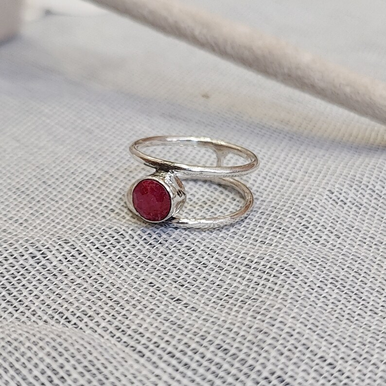 Może przedstawiać: Srebrny pierścionek z czerwonym kamieniem szlachetnym osadzonym w środku. Pierścionek ma podw&oacute;jny wz&oacute;r.