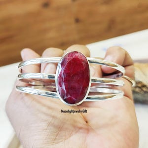 Puede incluir: Un brazalete de plata con tres bandas y una gran piedra preciosa de rubí rojo de forma ovalada en el centro. El brazalete está en una mano.