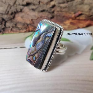 May include: A silver ring with a rectangular abalone shell inlay. The shell has iridescent colors of blue, green, and purple. The ring has a rope-like design around the edge.