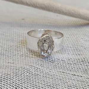Op de afbeelding: Een zilveren ring met een grote, heldere, gefacetteerde edelsteen in het midden. De ring is dik en heeft een eenvoudig, modern ontwerp.