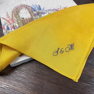 K&ouml;nnte beinhalten: Eine senfgelbe Stoffserviette mit dem in Dunkelblau gestickten Monogramm S & M in Schreibschrift. Die Serviette ist gefaltet und liegt auf einem hellen, rechteckigen Gegenstand, der mit einem bunten Blumen-Stickmuster verziert ist.