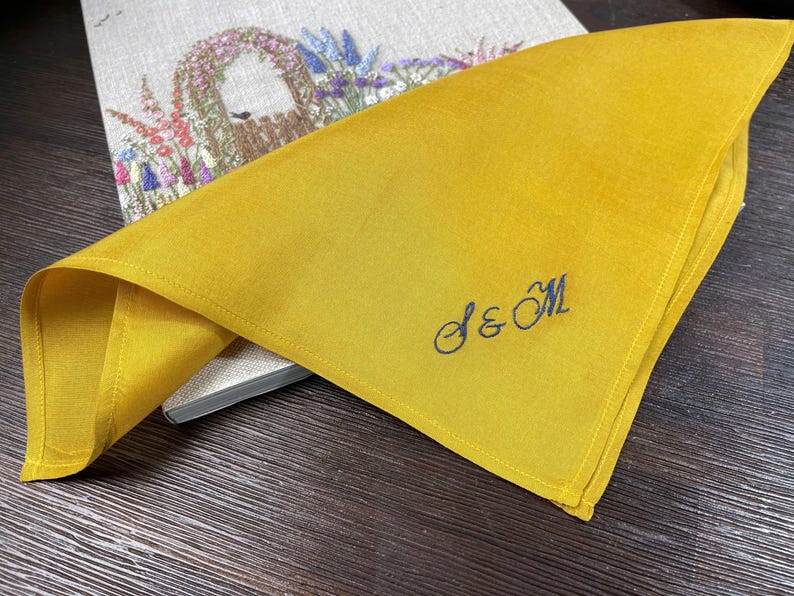 K&ouml;nnte beinhalten: Eine senfgelbe Stoffserviette mit dem in Dunkelblau gestickten Monogramm S & M in Schreibschrift. Die Serviette ist gefaltet und liegt auf einem hellen, rechteckigen Gegenstand, der mit einem bunten Blumen-Stickmuster verziert ist.