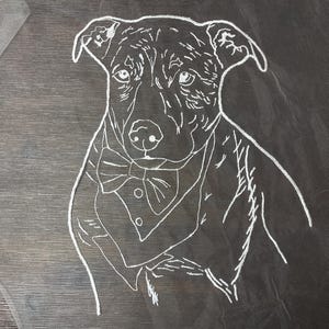 Pode incluir: Desenho a traço branco de um cão usando um smoking e uma gravata borboleta em tecido transparente. Os traços do cão são detalhados, com olhos, nariz e orelhas visíveis. O tecido está colocado sobre uma superfície de madeira escura.