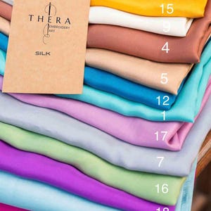 K&ouml;nnte beinhalten: Ein Stapel gefalteter Seidenstoffst&uuml;cke in verschiedenen Unifarben, darunter Pink, Gelb, Wei&szlig;, Braun, Beige, T&uuml;rkis, Lavendel, Hellgrau, Hellgr&uuml;n und Lila. Obenauf liegt eine beige Karte mit dem Text THERA EMBROIDERY ART SILK.