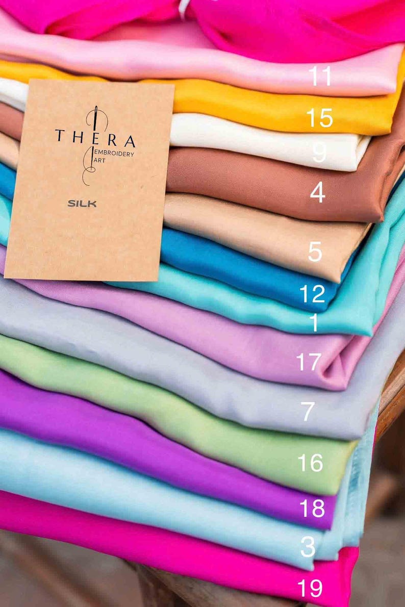 K&ouml;nnte beinhalten: Ein Stapel gefalteter Seidenstoffst&uuml;cke in verschiedenen Unifarben, darunter Pink, Gelb, Wei&szlig;, Braun, Beige, T&uuml;rkis, Lavendel, Hellgrau, Hellgr&uuml;n und Lila. Obenauf liegt eine beige Karte mit dem Text THERA EMBROIDERY ART SILK.