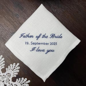 K&ouml;nnte beinhalten: Ein quadratisches wei&szlig;es Leinentaschentuch mit dunkelblauer Stickerei, die Father of the Bride, 19. September 2025, und I love you liest. Der Stoff hat eine sichtbare Webstruktur und ist schr&auml;g auf einer dunklen Oberfl&auml;che platziert.