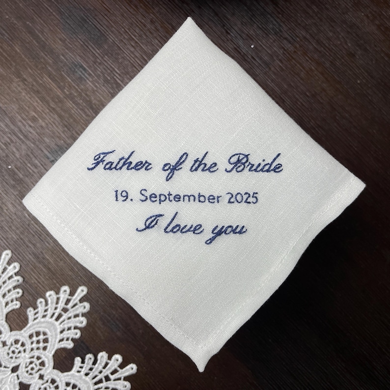 K&ouml;nnte beinhalten: Ein quadratisches wei&szlig;es Leinentaschentuch mit dunkelblauer Stickerei, die Father of the Bride, 19. September 2025, und I love you liest. Der Stoff hat eine sichtbare Webstruktur und ist schr&auml;g auf einer dunklen Oberfl&auml;che platziert.