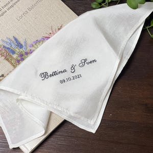 May include: A white fabric handkerchief featuring black embroidered text that reads Bettina & Sven and the date 09.10.2021. The handkerchief has a smooth texture and is folded to display the personalised lettering clearly on the fabric surface.