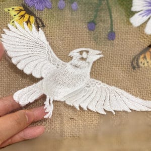 May include: A white bird embroidery patch on sheer mesh fabric, featuring detailed wing feathers. The background displays embroidered yellow and orange butterflies, purple floral buds, and white daisy-like flowers with yellow centers.