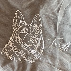 May include: A white embroidered line art portrait of a German Shepherd dog on a sheer fabric background. The embroidery features the name Trigger in a cursive script font next to a small embroidered heart outline, capturing the dog's facial features.