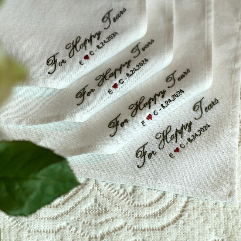 K&ouml;nnte beinhalten: Ein Stapel wei&szlig;er Stofftaschent&uuml;cher mit schwarzer Stickerei, die For Happy Tears liest, gefolgt von den Initialen E C und dem Datum 8.24.2024, mit einem kleinen roten Herz, das zwischen den Initialen und dem Datum eingestickt ist.