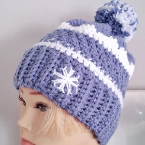 Könnte beinhalten: Eine blaue und weiße Häkelmütze mit einem weißen Bommel und einem weißen Schneeflockenmuster.