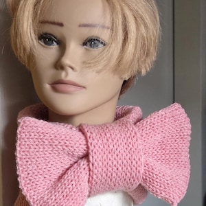 May include: A light pink, knitted bow scarf. The scarf is wrapped around a mannequin's neck, showcasing its bow-like design. The knitted texture and soft pink color give the scarf a cozy and feminine appearance. The scarf is a fashion accessory.