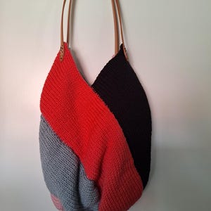Könnte beinhalten: Handgefertigte Tragetasche mit Farbblock-Design. Die Tasche hat Abschnitte aus rotem, schwarzem und grauem Strickstoff. Sie hat braune Lederriemen und hängt an einer weißen Wand. Der Text "sk.headbands" ist sichtbar.