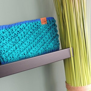 Könnte beinhalten: Handgefertigte Häkeltasche in Türkis und Blau. Die rechteckige Tasche hat ein strukturiertes Stichmuster und einen blauen Rand. Ein kleines braunes Etikett mit Text ist angebracht. Die Tasche wird auf einem dunkelgrauen Regal präsentiert.