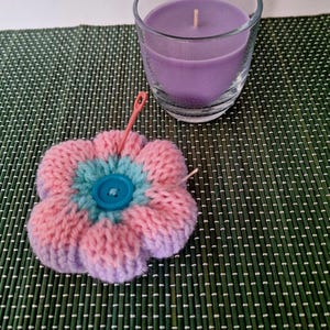 Könnte beinhalten: Eine gestrickte Blume in Pink und Lila mit einem blauen Knopf in der Mitte. Die Blume liegt auf einer grünen gewebten Matte mit einem Glasgefäß mit einer lila Kerze im Hintergrund.