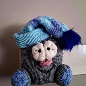 Könnte beinhalten: Ein graues, gestricktes Plüschtier mit einer blauen und violetten Strickmütze. Das Spielzeug hat ein lächelndes Gesicht mit großen weißen Augen, einer herausgestreckten rosa Zunge und blauen Strickfüßen.